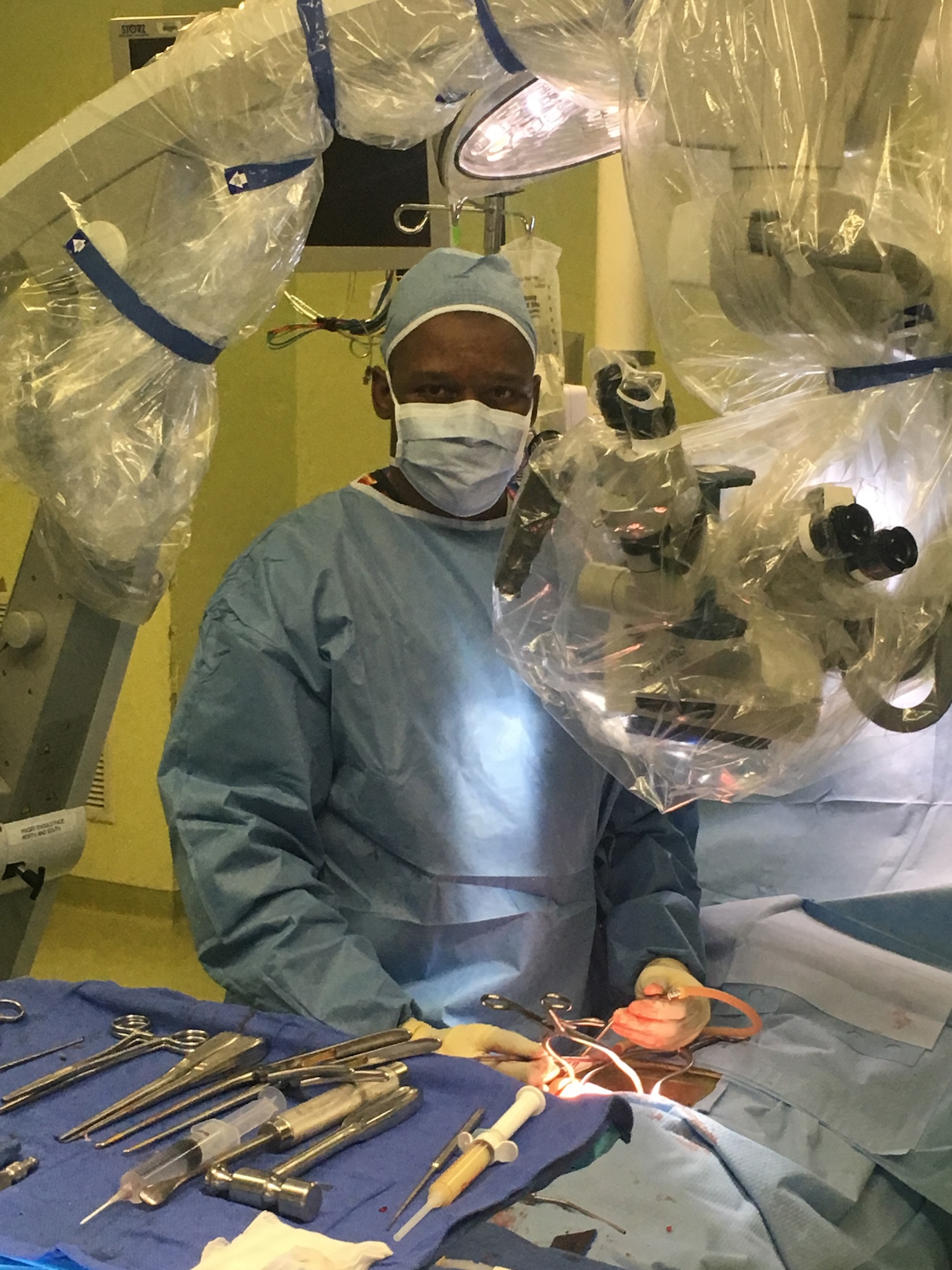 A person wearing medical scrubs and a surgical mask stands behind a table with various medical equipment and supplies, including a large microscope with an illuminated stage and a magnifying glass, set up within a hospital operating room environment.