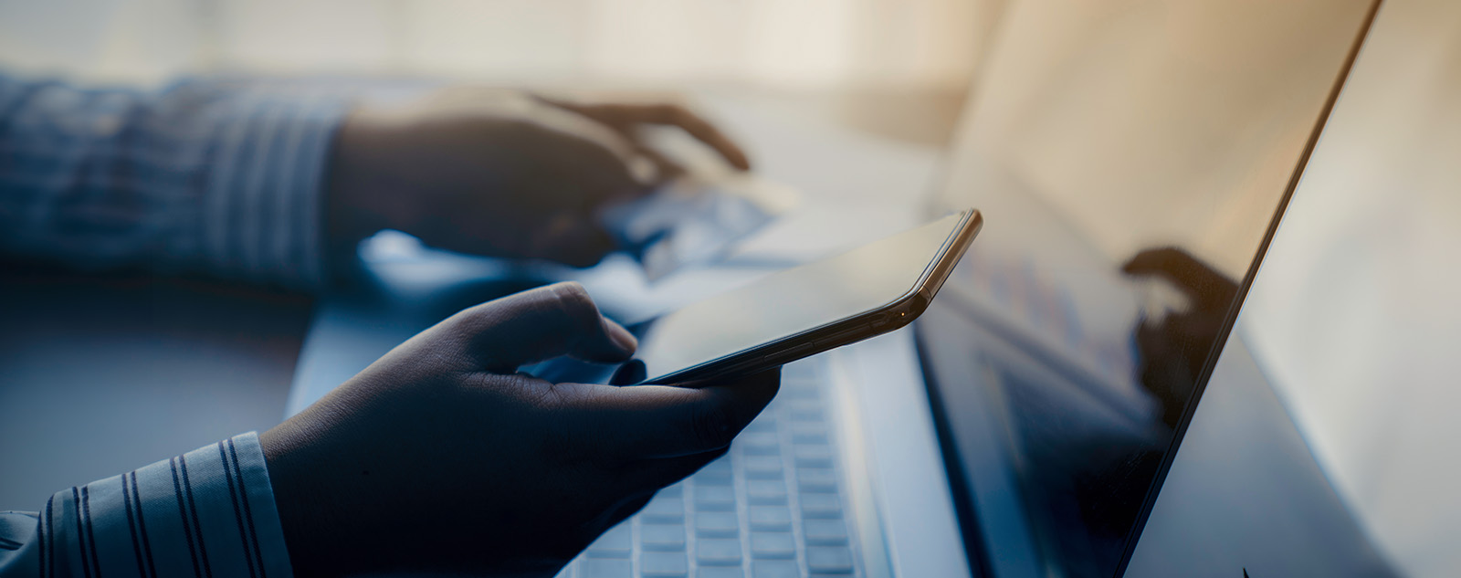 The image shows a person s hands working on a laptop computer with a blurred background featuring a smartphone and a table, suggesting multitasking or productivity.