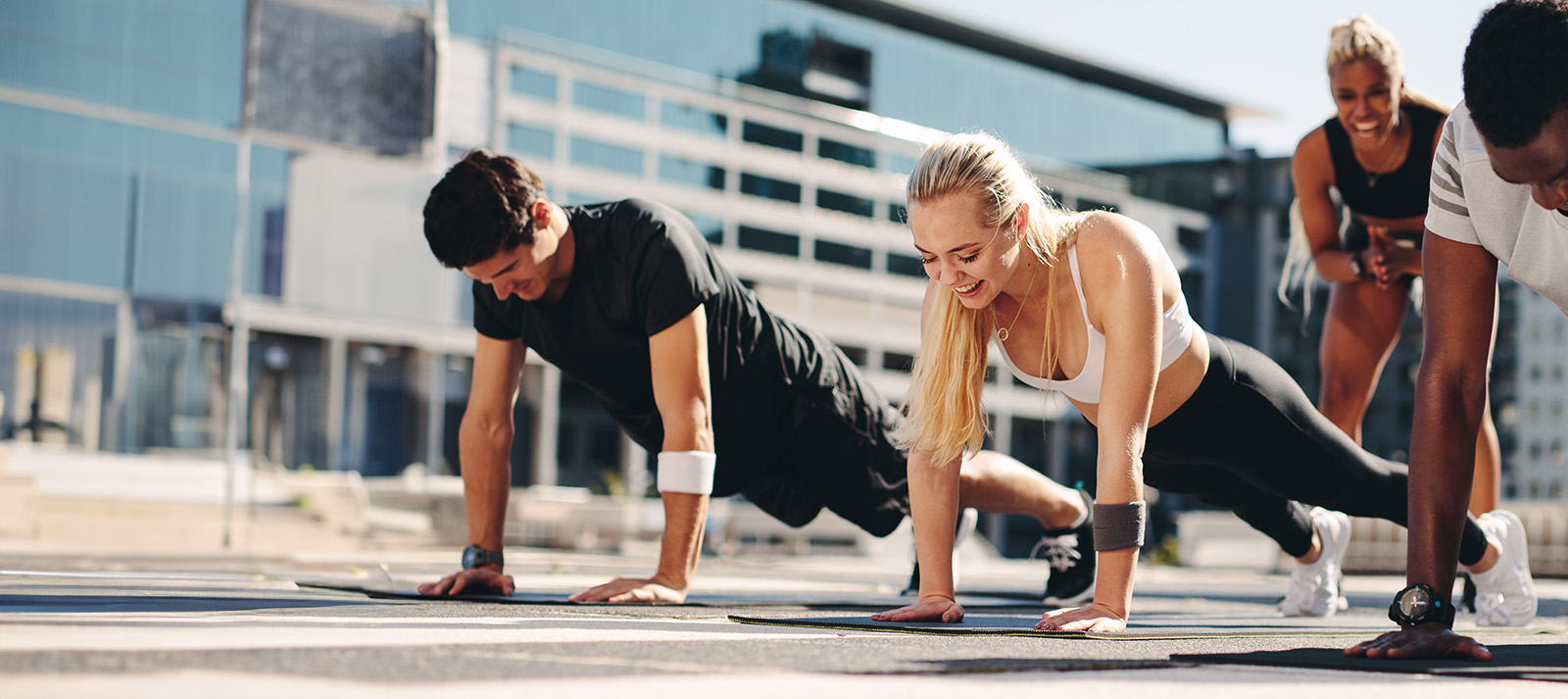 A group of people performing a workout exercise with their hands on the ground and legs lifted in the air, set against an urban background.