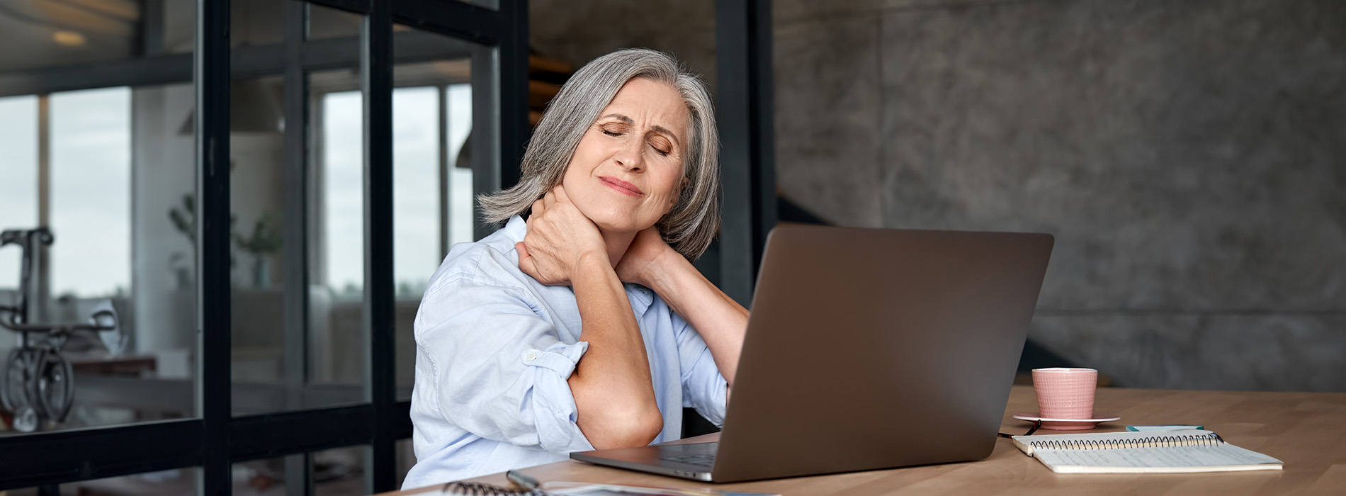 The image shows a woman seated at a table with a laptop in front of her, leaning on her hand with her elbow resting on the table, looking off to the side with a thoughtful expression.