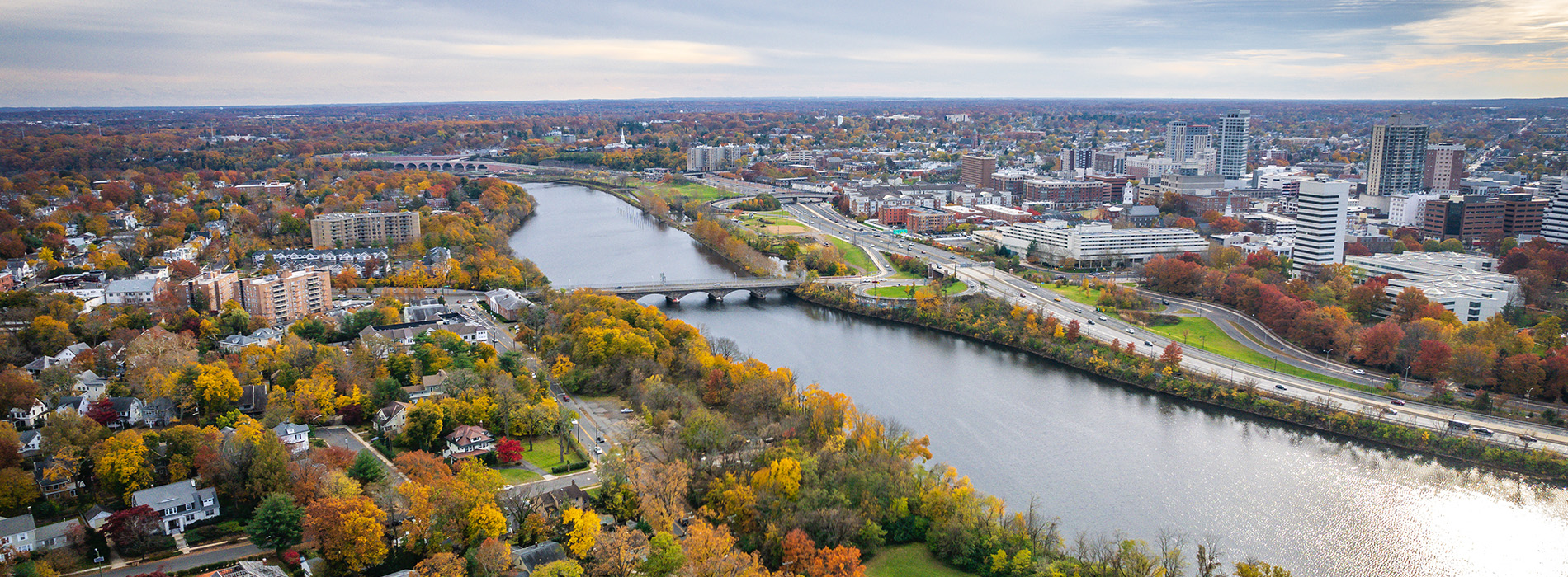 The image shows a scenic view of a city skyline with a river running through it, surrounded by autumn foliage, under a partly cloudy sky.