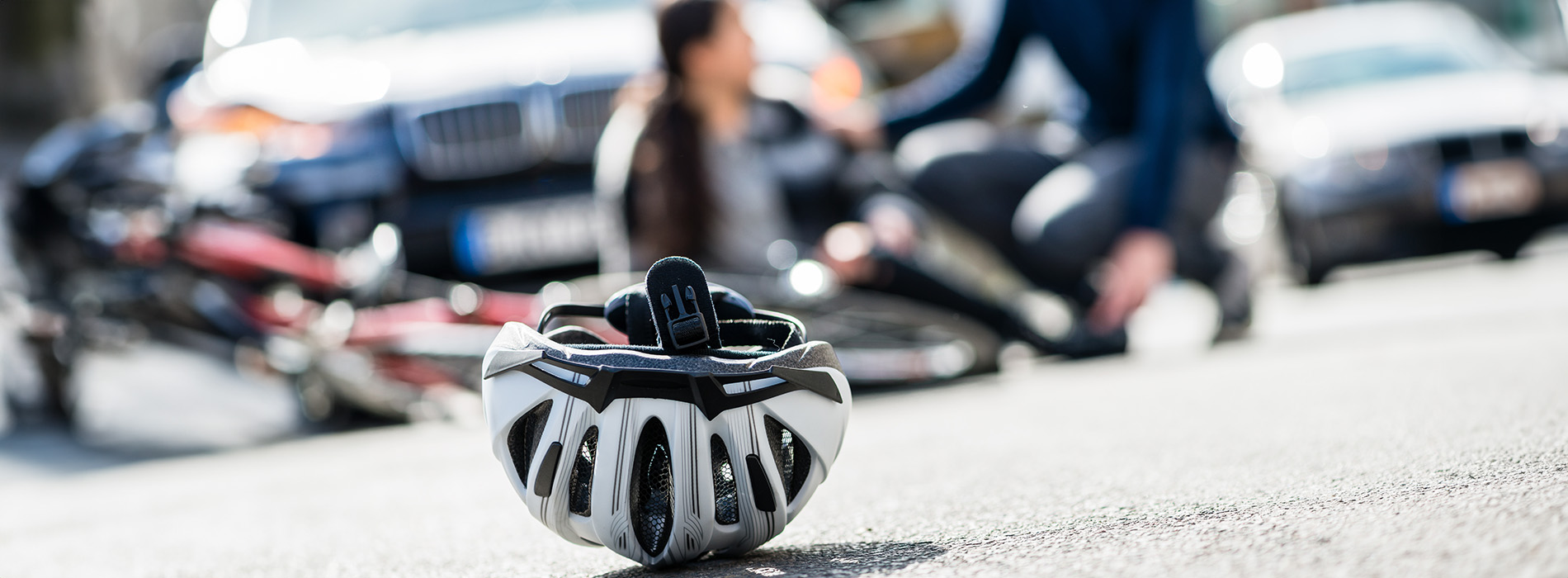 The image shows a person on a bicycle with a helmet, positioned against a blurred background of parked cars and a city street.