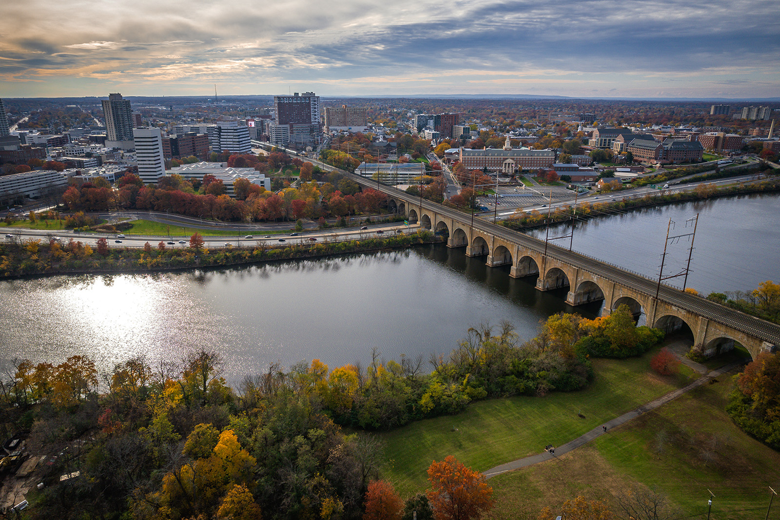 The image shows a picturesque autumnal scene featuring a river with a bridge over it, surrounded by trees with fall colors, set against a city skyline under an overcast sky.
