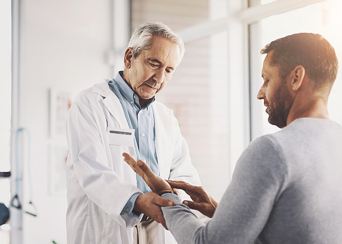 The image depicts a man in a white coat assisting another man with his hand, which appears to be injured or in need of attention. They are in an indoor setting that looks like a medical office.