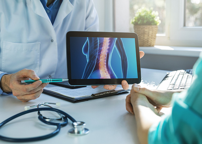 The image depicts a medical professional using a tablet with an X-ray image displayed, showing a spine, while another person observes the screen.