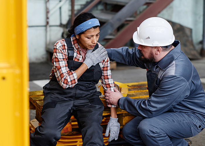 The image shows a man in protective gear attending to a woman who appears to be experiencing discomfort or pain  they are in an industrial setting with machinery in the background.