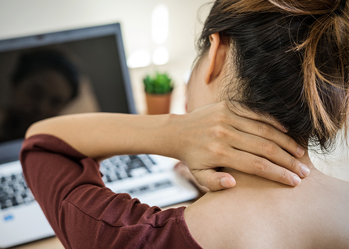 A woman with her arm resting on a laptop screen, massaging her neck while seated at a desk.
