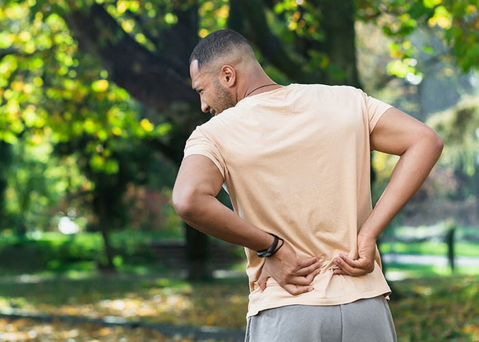 The image features a man standing outdoors, wearing a light-colored t-shirt, with his back turned to the camera. He appears to be looking over his shoulder, possibly at something off-camera, and has one hand placed on his lower back.